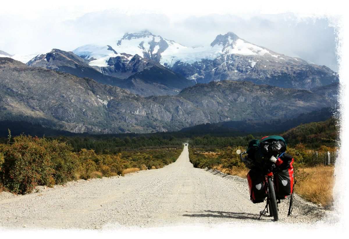 A llong gravel road with snow cover mountains at the horizon. A bikepacking bike near the side of the road.